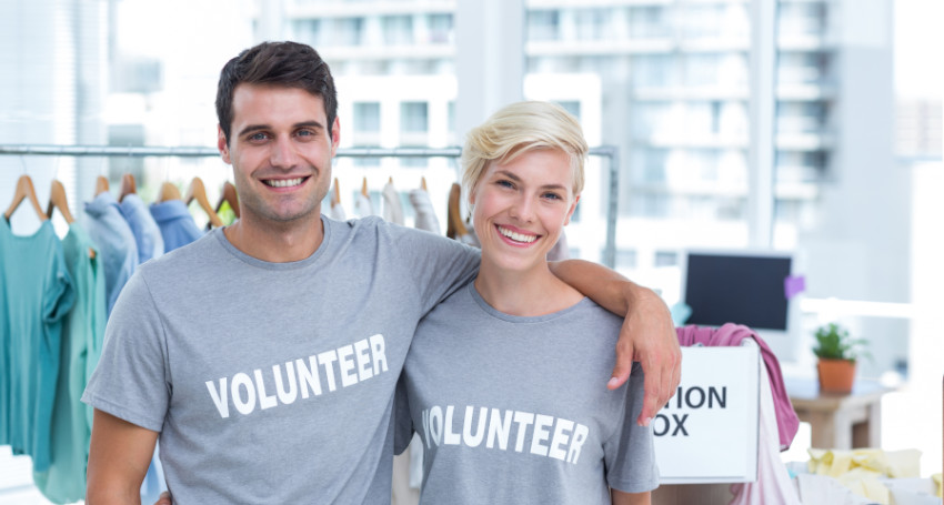 Two cheerful volunteers wearing gray t-shirts labeled "volunteer" standing in a clothing donation center.
