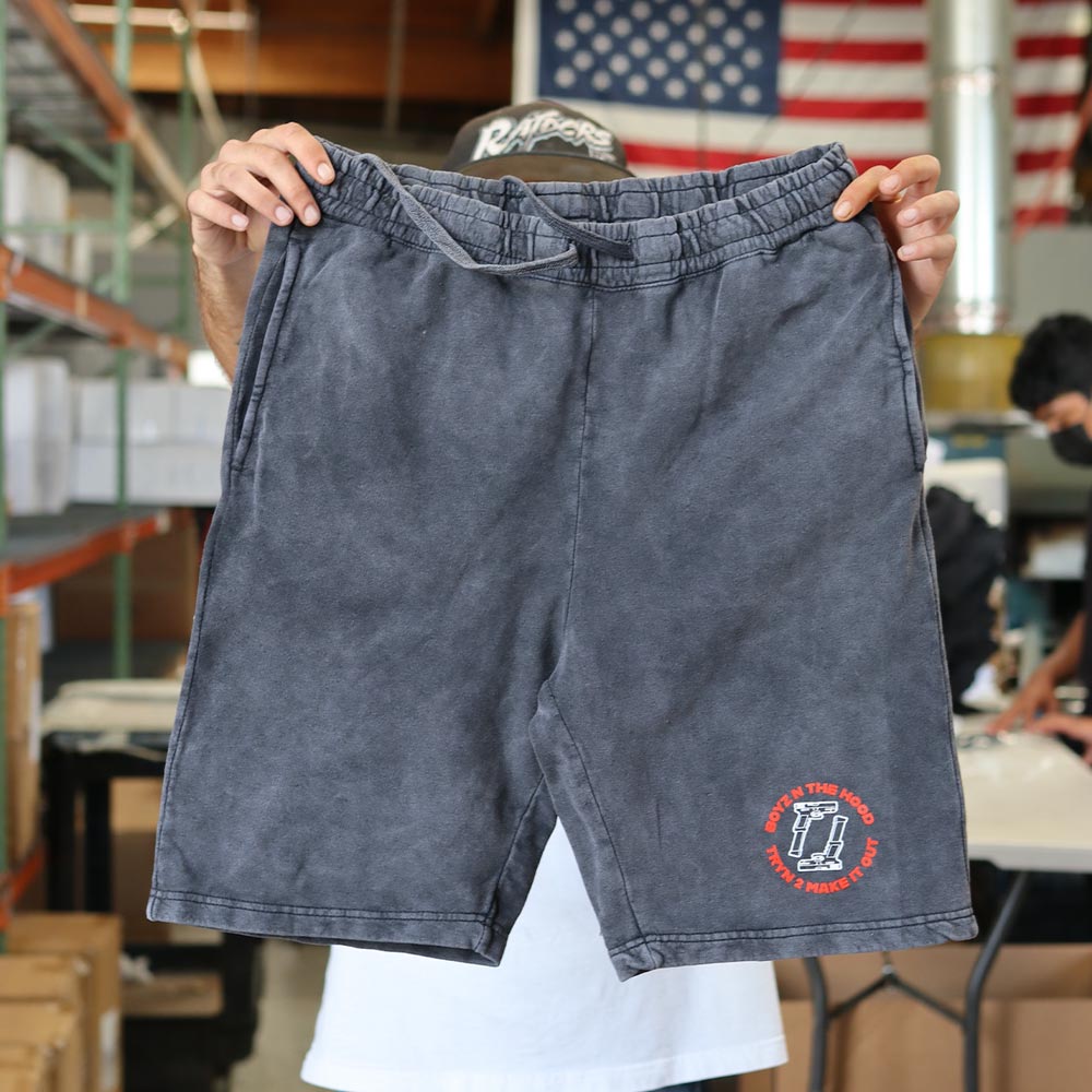 A man holding the front view of a Mineral Wash Black Independent Trading Co. PRM50STMW MENS MINERAL WASH FLEECE SHORT featuring the custom screen print of "Boyz n the hood tryn 2 make it out" in bold orange text arranged in a circular format with 2 guns inside printed by Garment Decor