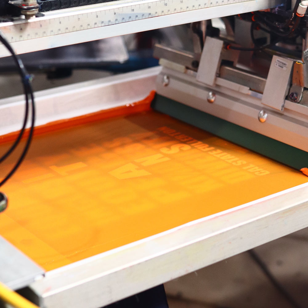 Capturing the screen printing process, this image shows the Titans Cal State Fullerton design being expertly printed on a Gildan Hammer™ T-Shirt using an M&R Print Machine printed by Garment Decor. Advanced technology ensures accuracy and vibrant results.