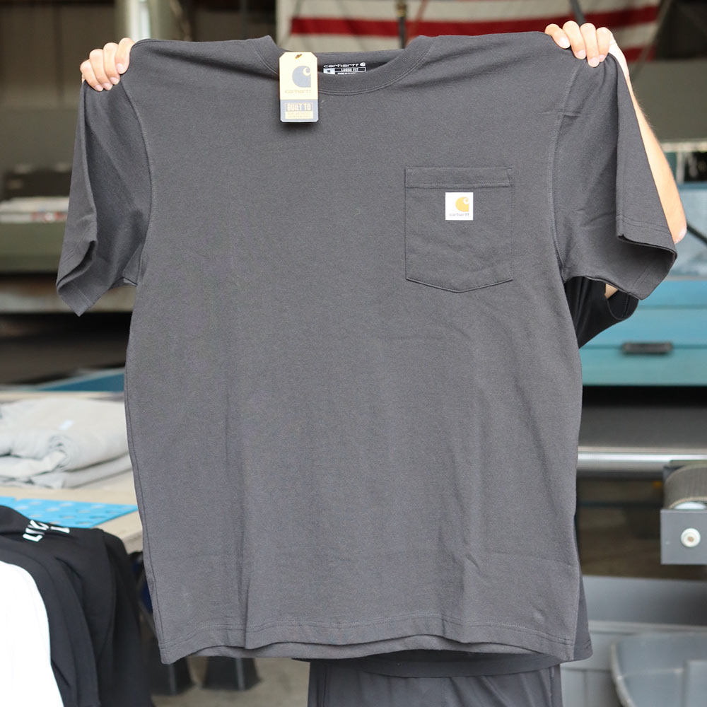A man holding the front of a Carhartt® CTK87 Workwear Pocket Short Sleeve T-Shirt in Bluestone, showcasing the left chest pocket with Carhartt® woven label