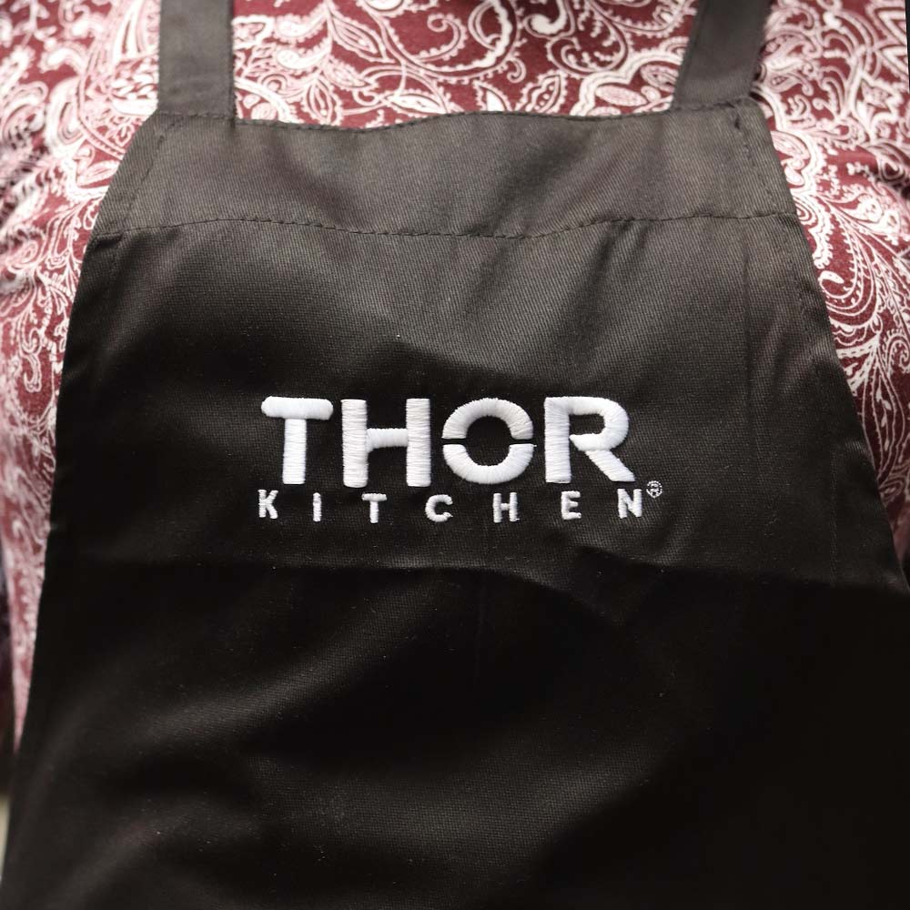 The photo focuses on the intricate "Thor Kitchen" embroidery on the Port Authority® A700 Easy Care Extra Long Bib Apron in black. The design highlights the high-quality embroidery work by Garment Decor, ensuring durability and precision.
