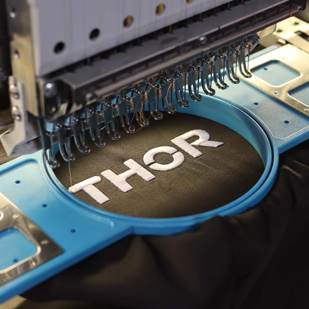 This photo captures the production process of the black Port Authority® A700 Easy Care Extra Long Bib Apron as the "Thor Kitchen" design is embroidered on a Barudan embroidery machine. Garment Decor's expertise ensures a flawless result for every piece.