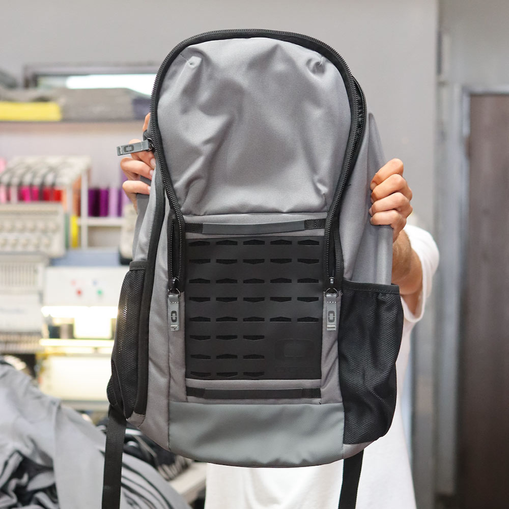 This front view of gray and black backpack stands out with its custom "DFX4 Victorville, CA" logo embroidered by Garment Decor, showcasing fine detail and craftsmanship. The bag features a sleek design with mesh side pockets for extra storage and a durable, structured front panel, making it both stylish and practical for daily use or special events. Perfect for organizations, teams, or branded giveaways.