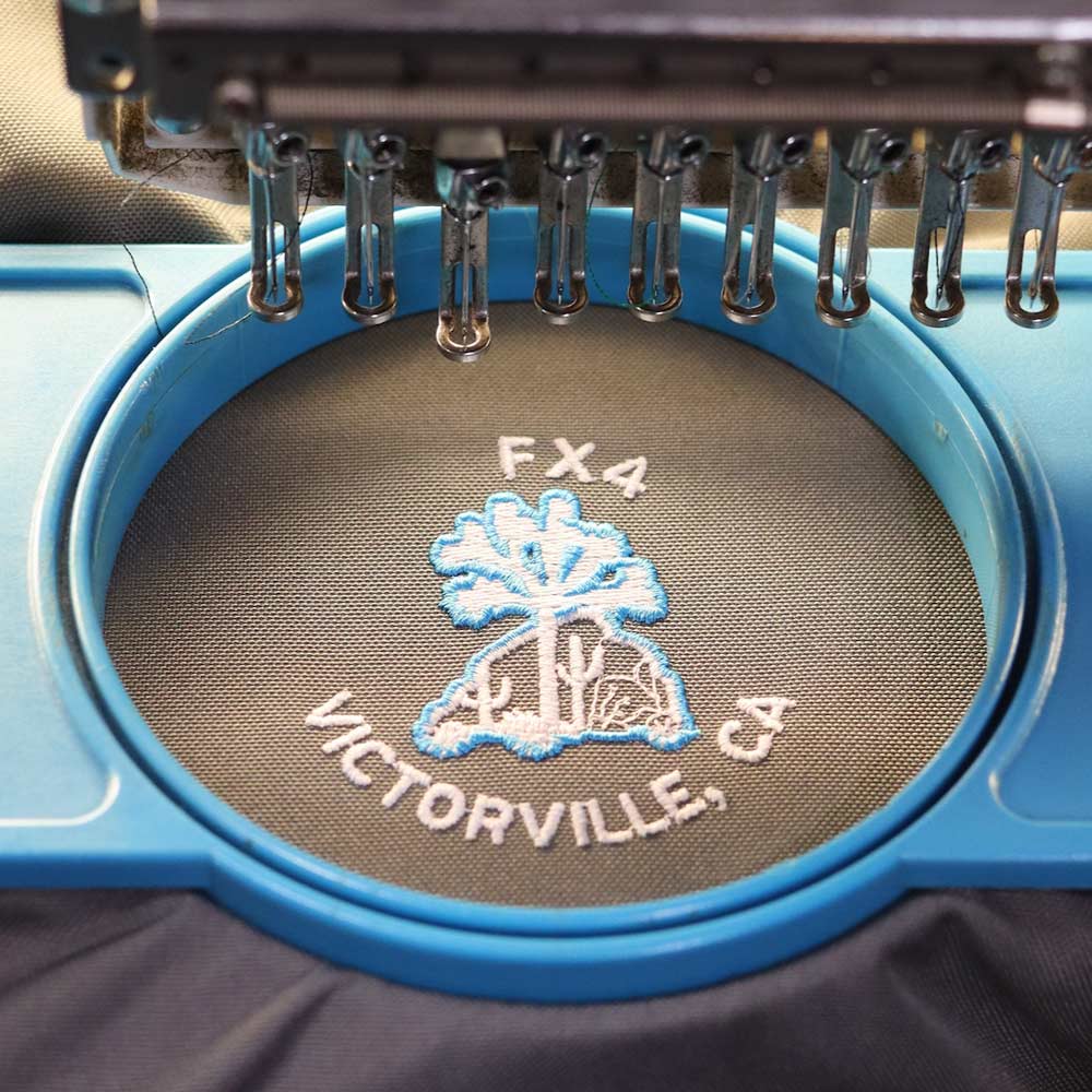 This front view of gray and black backpack while being embroidered under a Barudan machine stands out with its custom "DFX4 Victorville, CA" logo embroidered by Garment Decor, showcasing fine detail and craftsmanship. The bag features a sleek design with mesh side pockets for extra storage and a durable, structured front panel, making it both stylish and practical for daily use or special events. Perfect for organizations, teams, or branded giveaways.