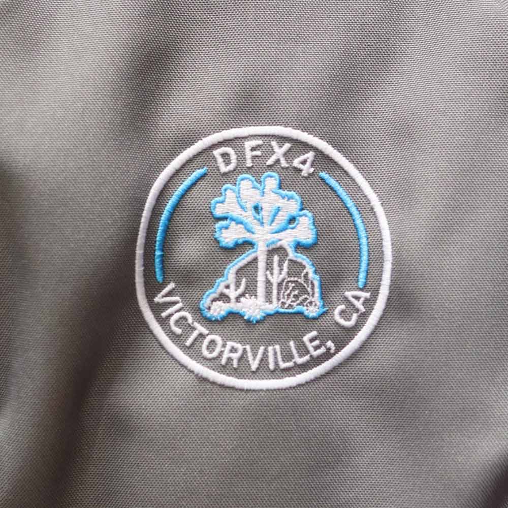This zoomed in front view of gray and black backpack stands out with its custom "DFX4 Victorville, CA" logo embroidered by Garment Decor, showcasing fine detail and craftsmanship. The bag features a sleek design with mesh side pockets for extra storage and a durable, structured front panel, making it both stylish and practical for daily use or special events. Perfect for organizations, teams, or branded giveaways.
