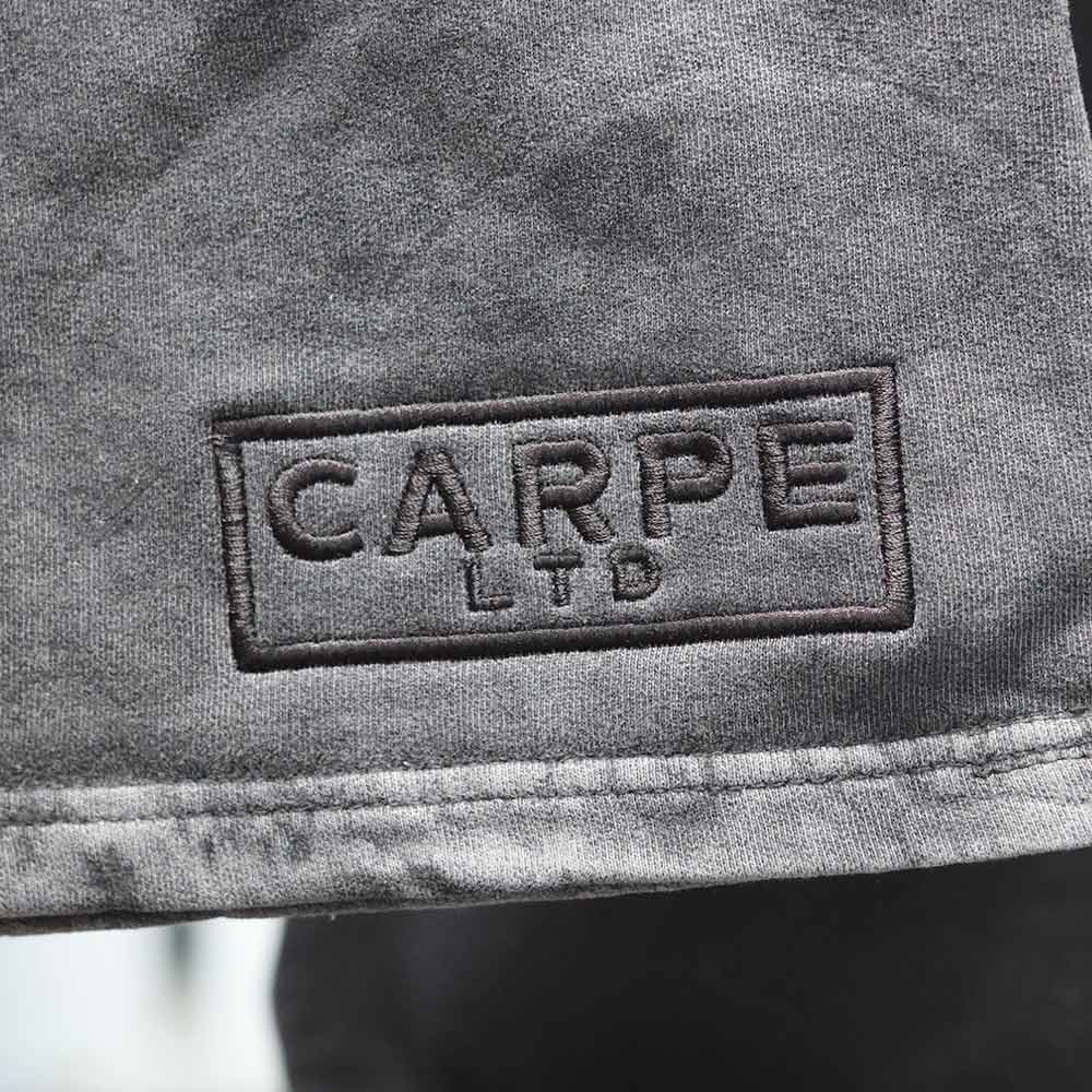 This close-up image captures the intricate details of the custom tonal 3D puff embroidery featuring the Carpe LTD design on the front view of the Independent Trading Co. PRM50STMW Mineral Wash Fleece Shorts in black. Embroidered by Garment Decor, the zoomed-in view emphasizes the precision and craftsmanship involved in creating this bespoke piece.