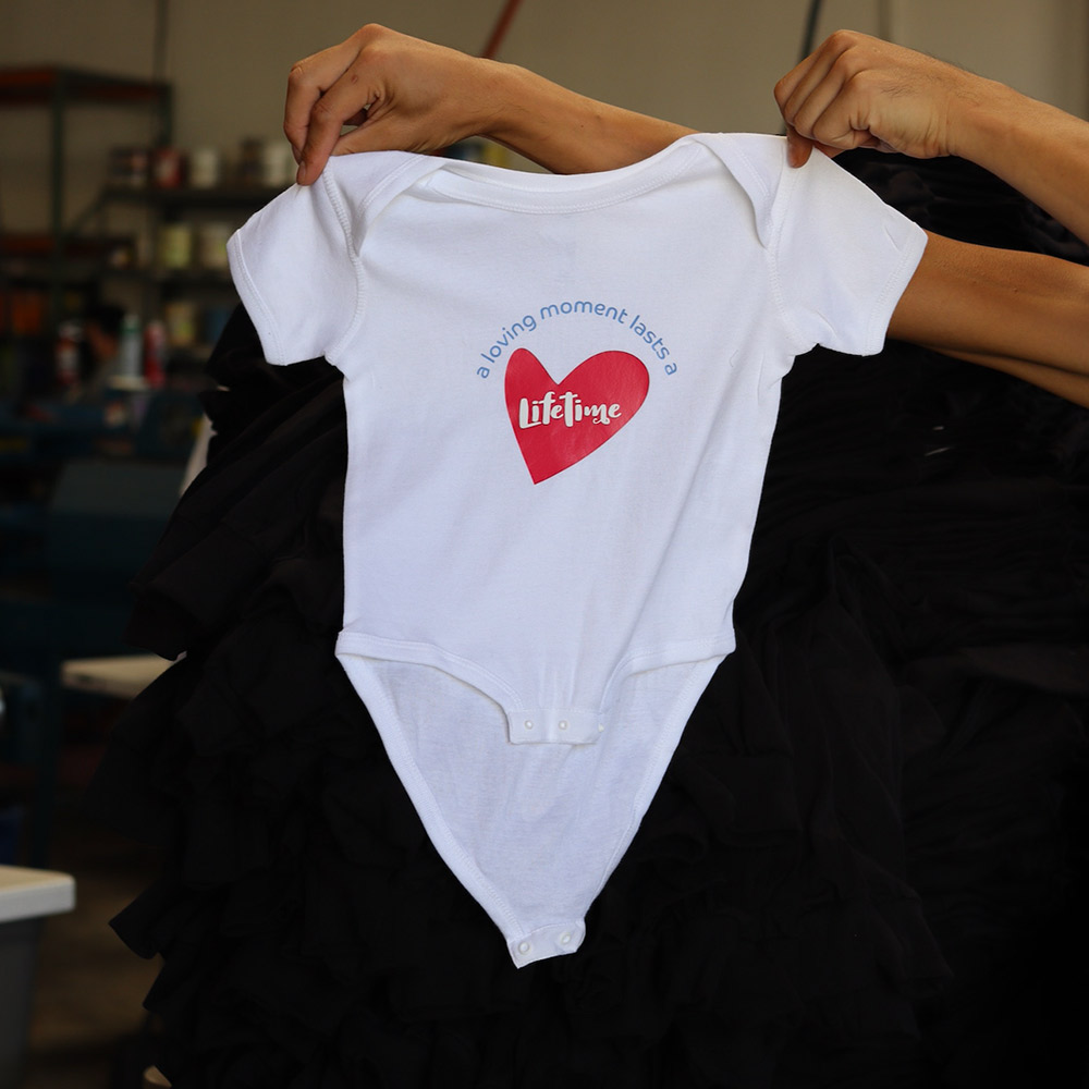A man holding this Rabbit Skins 4400 Infant Baby Rib Bodysuit in white combines softness and practicality for infants. The front showcases the sentimental "a loving moment lasts a lifetime" design, applied with precision using custom transfer techniques by Garment Decor. Perfect for baby gifts, special occasions, or everyday use, this premium bib ensures both comfort and style for your little one.