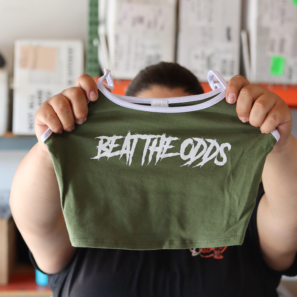 A close-up of a person holding a military green Mira Luxe ML-MTP024 tank top featuring a bold white "BEAT THE ODDS" design, custom screen-printed by Garment Decor.