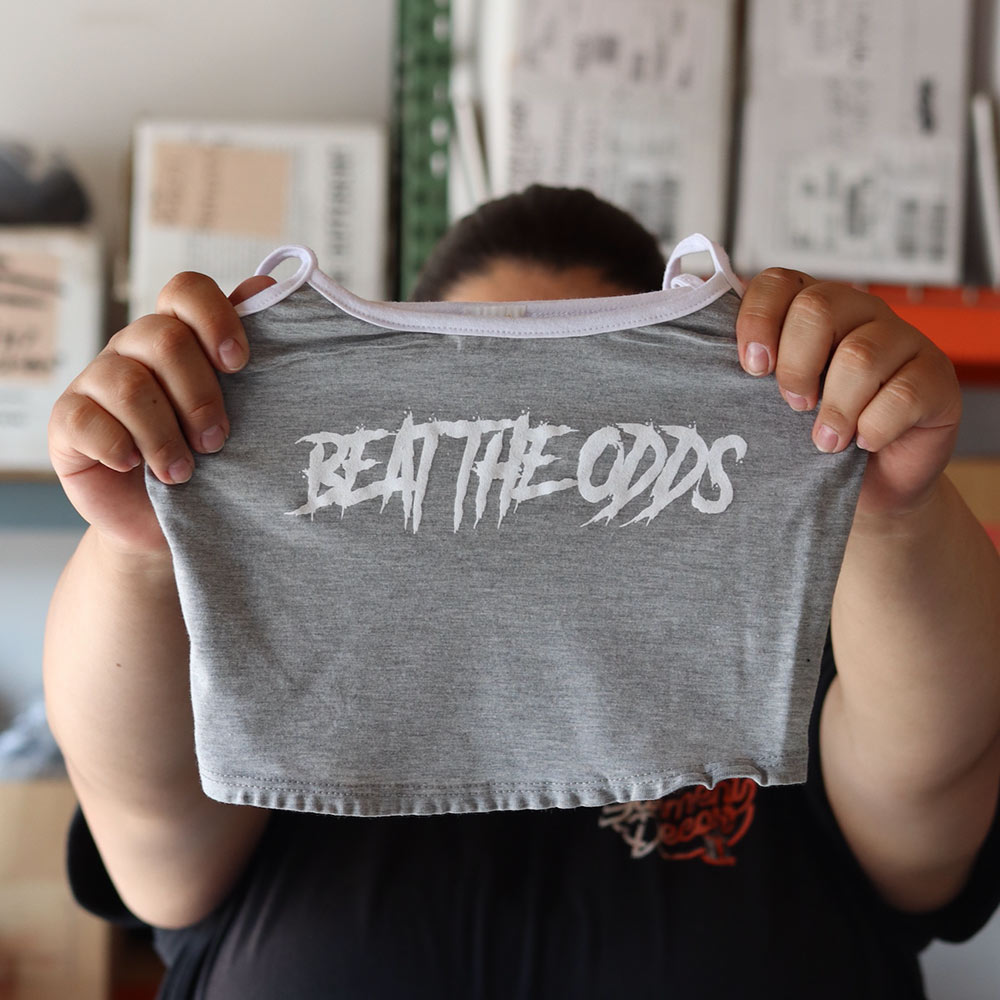 A close-up of a person holding a grey Mira Luxe ML-MTP024 tank top featuring a bold white "BEAT THE ODDS" design, custom screen-printed by Garment Decor.