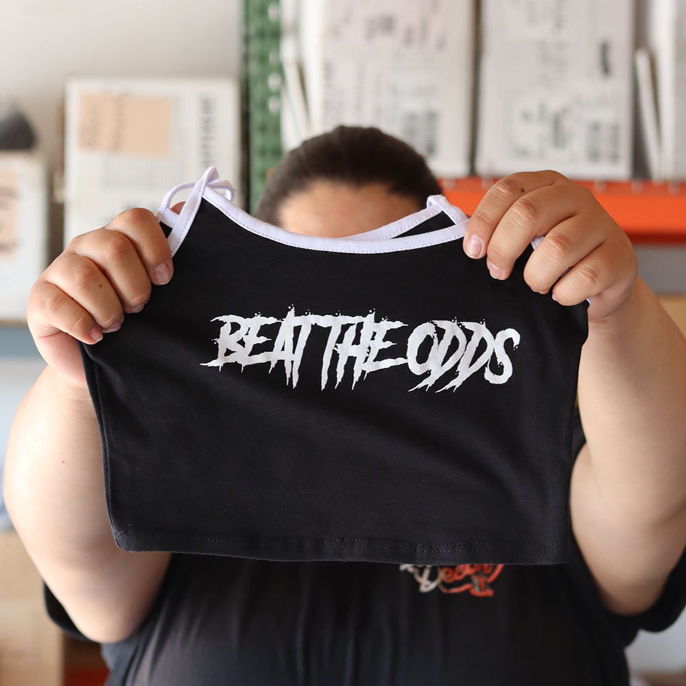 A close-up of a person holding a black Mira Luxe ML-MTP024 tank top featuring a bold white "BEAT THE ODDS" design, custom screen-printed by Garment Decor.