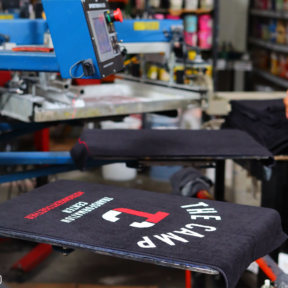 This image showcases the production process of Garment Decor's custom-printed towel for "The Camp Transformation Center." The black towel, featuring white and red text and logo, is positioned on a heat press machine in a busy workshop. The workspace includes printing equipment, supplies, and stacks of towels in the background. The vibrant and precise design reflects high-quality printing, ideal for branding and promotional materials.