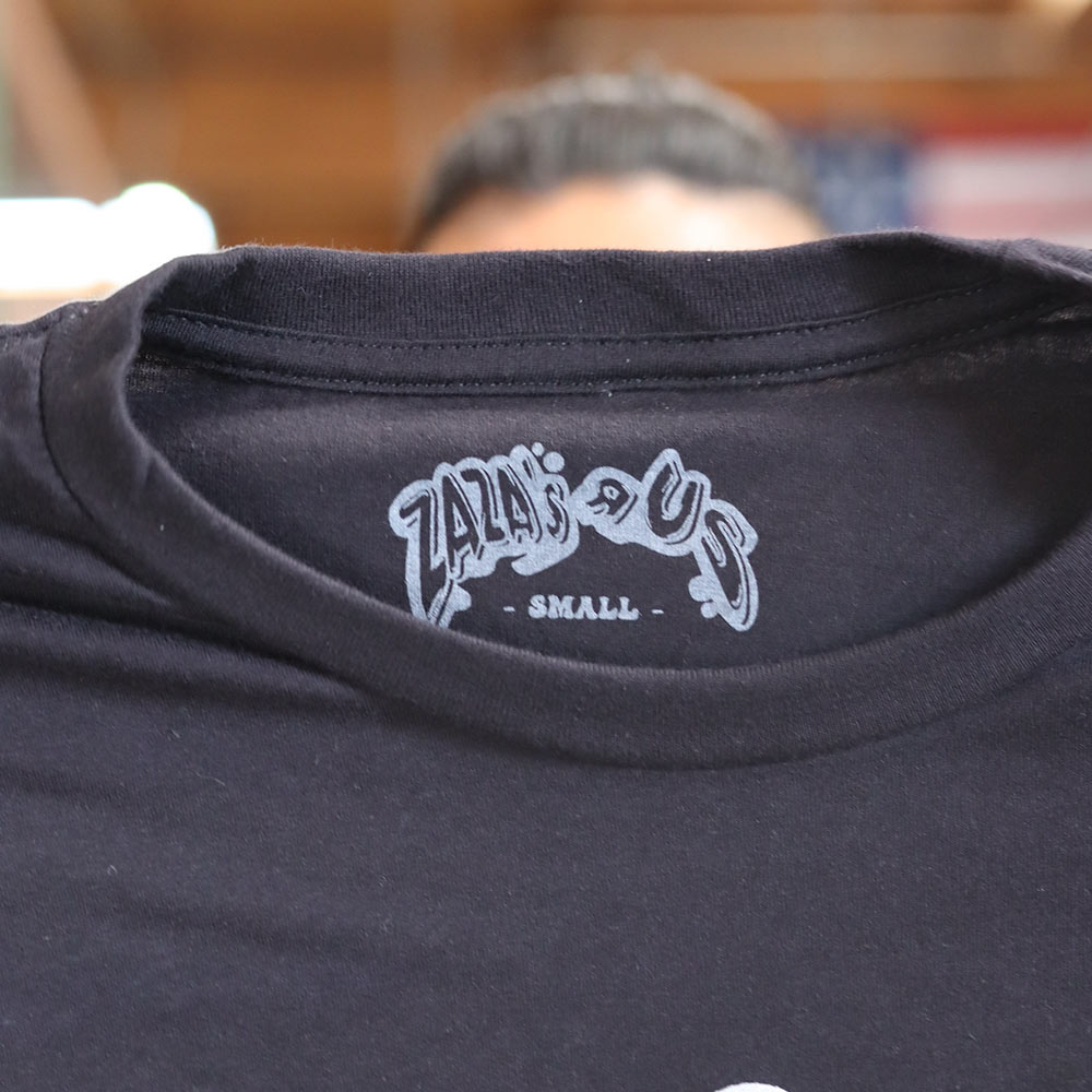 A man holding the neck label view of a black BELLA + CANVAS 3001 Jersey t-shirt featuring the custom digital squeegee print of "Zaza's R US" logo in bold and vibrant colorful design printed by Garment Decor