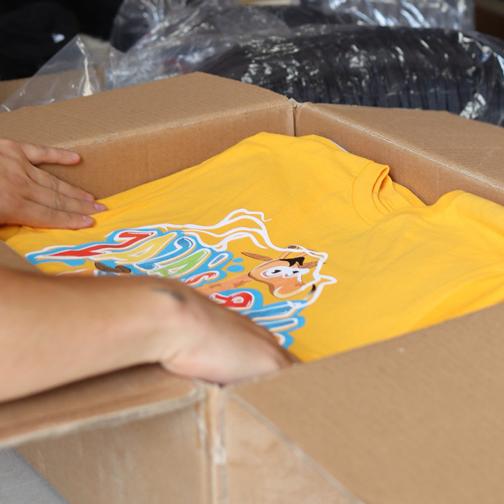 A man holding the front view of a yellow BELLA + CANVAS 3001 Jersey t-shirt featuring the custom digital squeegee print of "Zaza's R US" logo in bold and vibrant colorful design printed by Garment Decor while putting it inside the packaging box