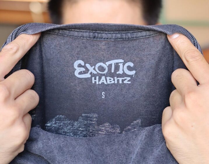 This photo showcases a man holding the neck label view of a Lane Seven LST002 5.5 oz Vintage Crewneck Tee in Vintage Black emphasizing the attention to detail in branding and garment size. The vibrant "Exotic Habitz" label was printed by Garment Decor, this tee combines unique artistry with top-quality printing.