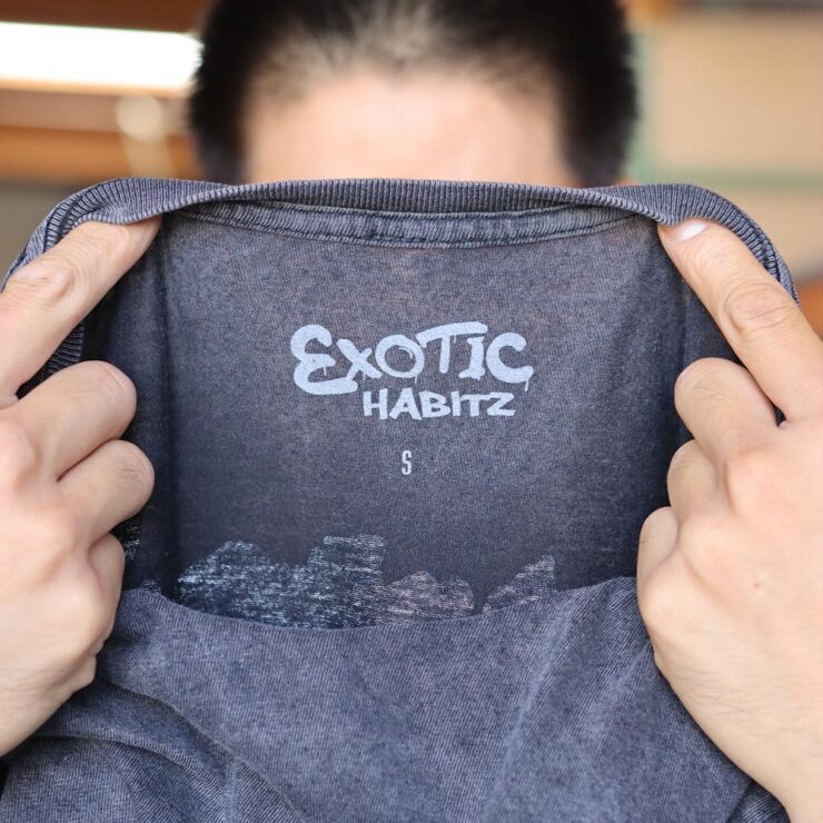 This photo showcases a man holding the neck label view of a Lane Seven LST002 5.5 oz Vintage Crewneck Tee in Vintage Black emphasizing the attention to detail in branding and garment size. The vibrant "Exotic Habitz" label was printed by Garment Decor, this tee combines unique artistry with top-quality printing.