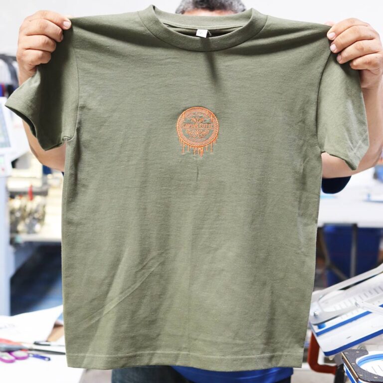 A person holding up a green t-shirt. This image highlights the front view of the AA 1301 Heavyweight Cotton Military Green T-Shirt custom embroidered for Caviar Cutz. Made with durable cotton, this shirt features intricate embroidery that elevates its premium look, perfect for professional branding.