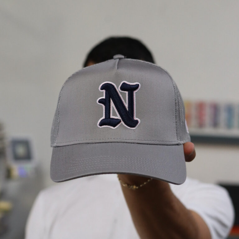 A man holding the front view of this cap. Elevate your brand with custom 3D puff embroidered Ottocap Style 32-285 trucker hat in Grey, featuring bold, raised designs that add texture and dimension to any logo or artwork. With a classic trucker style and breathable mesh back, these hats offer both comfort and standout style, making them a perfect choice for promoting your business or creating unique, fashionable headwear.