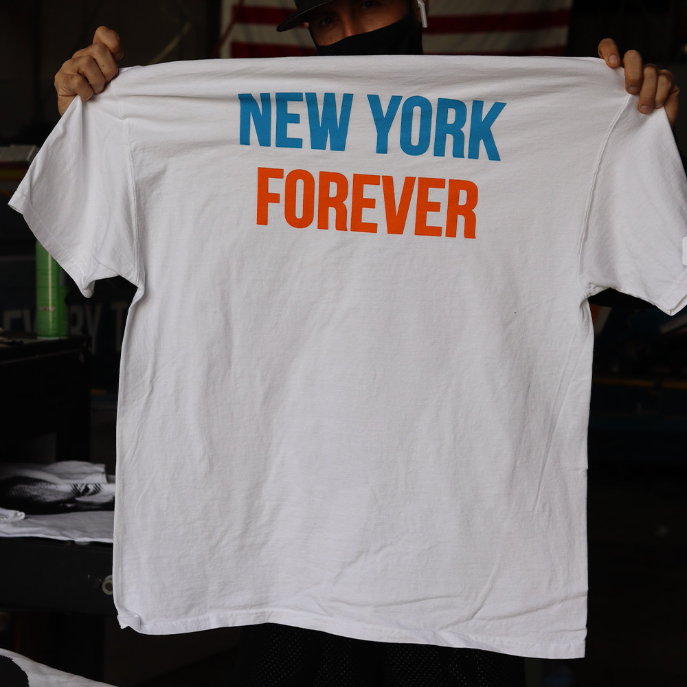 This photo showcases a man holding the back of a white Hanes GDH100 T-shirt featuring a custom design by Shmeel. The design prominently displays "New York" in blue and "Forever" in orange, creating a vibrant and eye-catching aesthetic. Screen printed by Garment Decor, this high-quality piece exemplifies precision printing and design creativity, making it a perfect addition for New York fans or custom apparel enthusiasts.