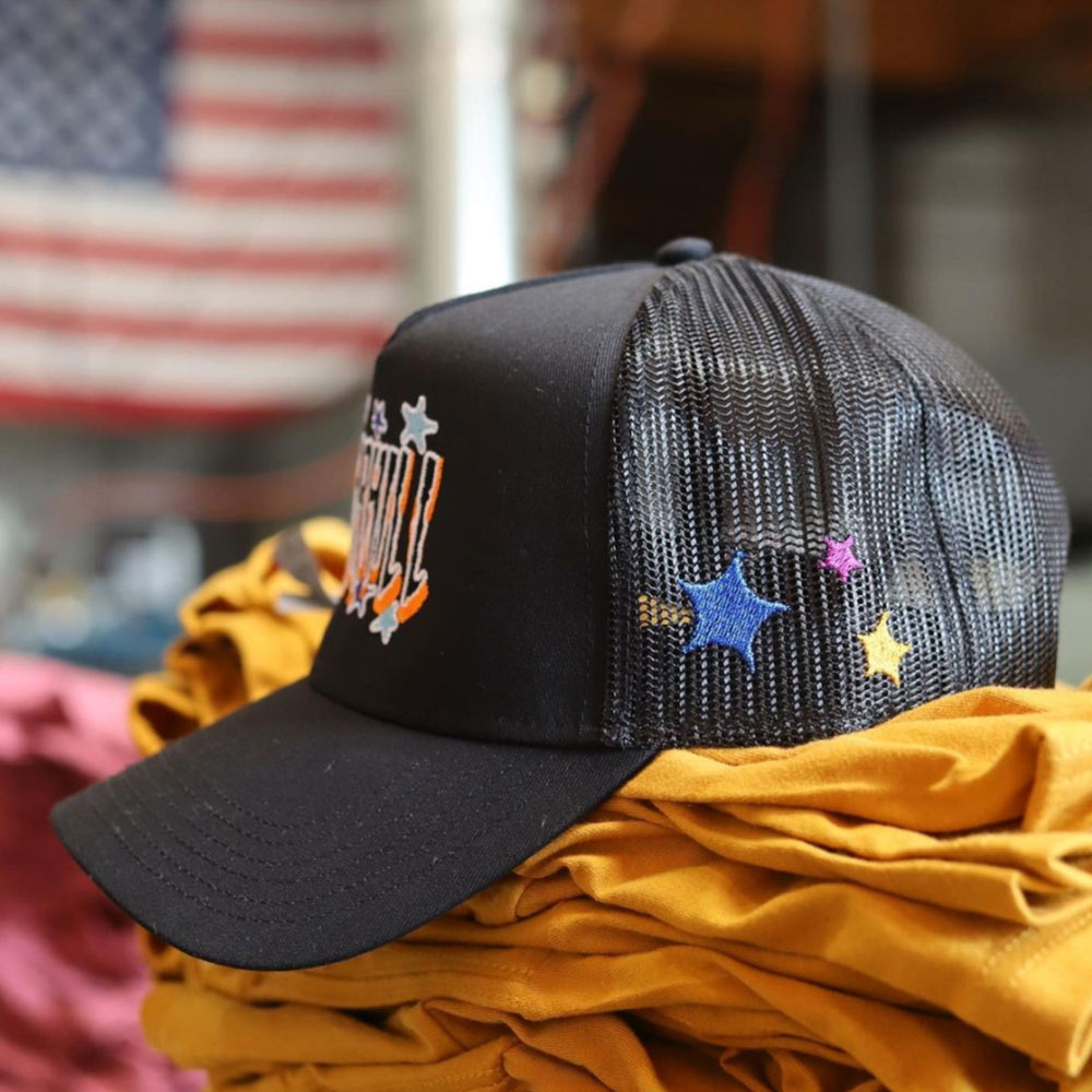 Black baseball cap with an emblem and colorful star designs on a yellow fabric surface, with an out-of-focus american flag in the background.