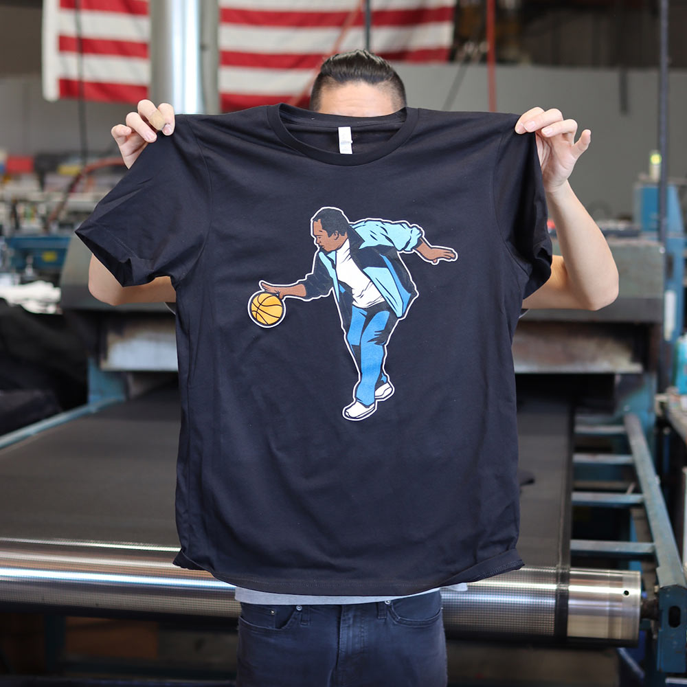 A sleek Bella + Canvas 3001 Unisex Jersey T-Shirt in black, featuring a custom screen print of The Baller design inspired by The Office merchandise. A man holds the shirt, showcasing the vibrant and detailed front print crafted by Garment Decor, perfect for fans and collectors alike.