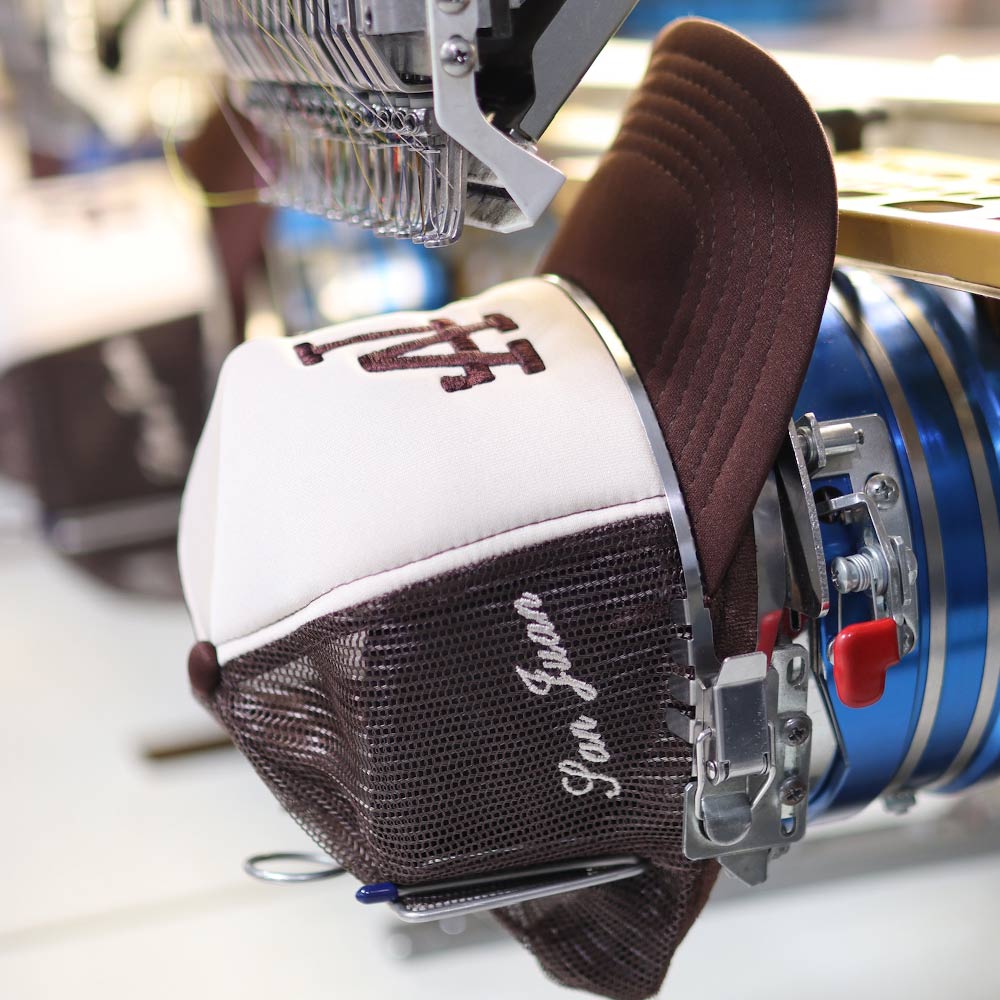 An up-close side view of the OTTO CAP 32-467 5-Panel Mid Profile Mesh Back Trucker Hat in Brown/Tan/Brown being embroidered with precision under a Barudan machine. The LA design comes to life with Garment Decor's custom embroidery expertise, highlighting quality craftsmanship.