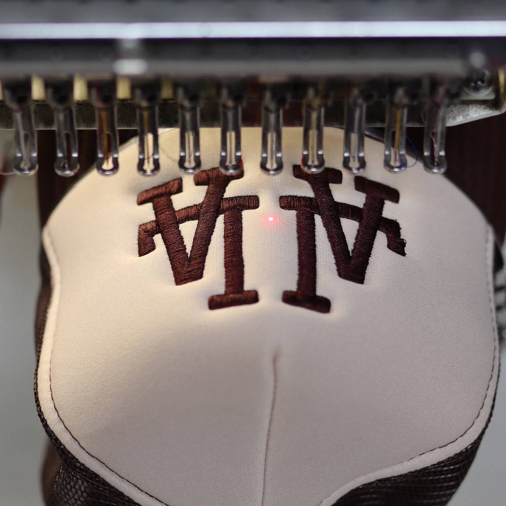 An up-close front view of the OTTO CAP 32-467 5-Panel Mid Profile Mesh Back Trucker Hat in Brown/Tan/Brown being embroidered with precision under a Barudan machine. The LA design comes to life with Garment Decor's custom embroidery expertise, highlighting quality craftsmanship.