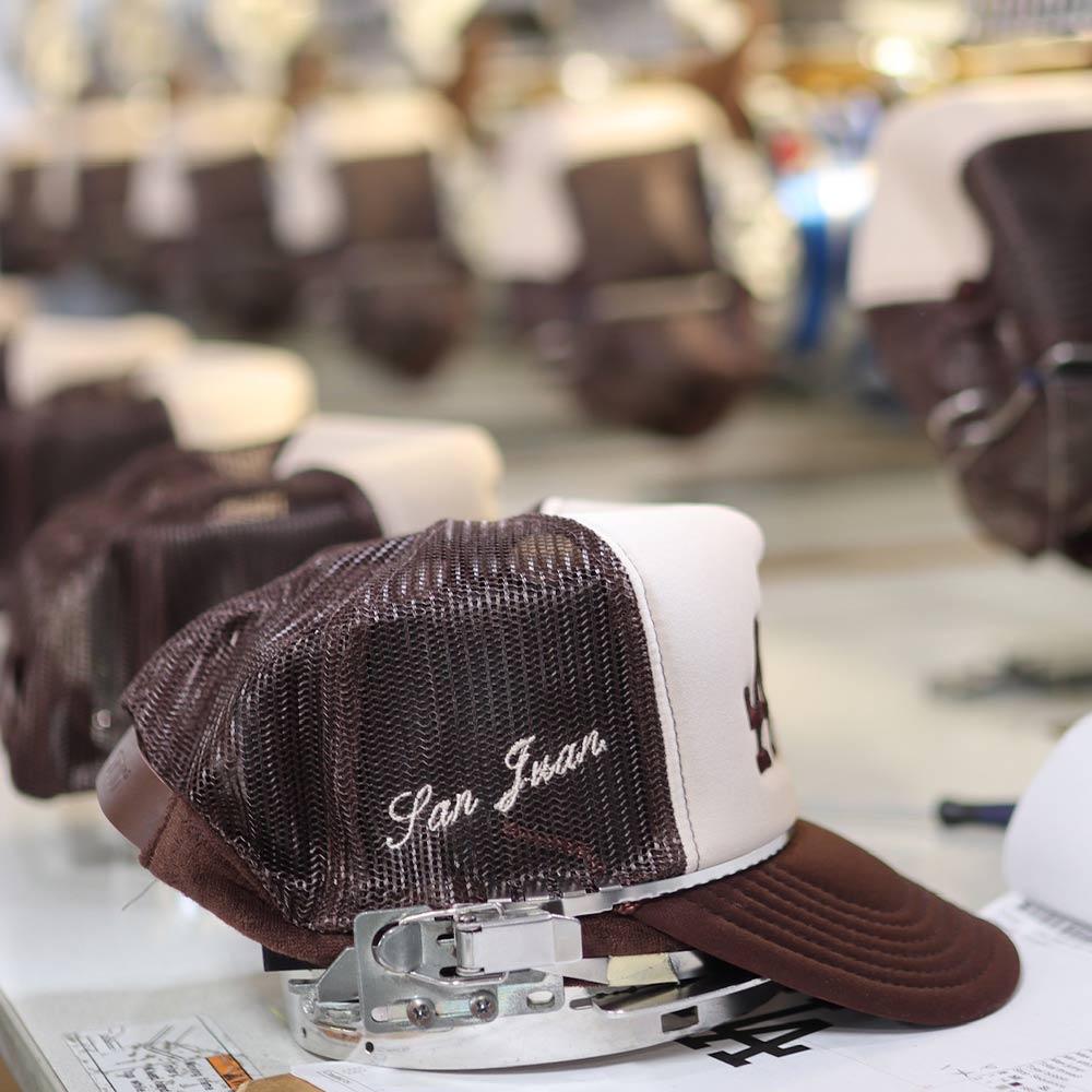 An up-close side view of the OTTO CAP 32-467 5-Panel Mid Profile Mesh Back Trucker Hat in Brown/Tan/Brown being embroidered with precision under a Barudan machine. The LA design comes to life with Garment Decor's custom embroidery expertise, highlighting quality craftsmanship.