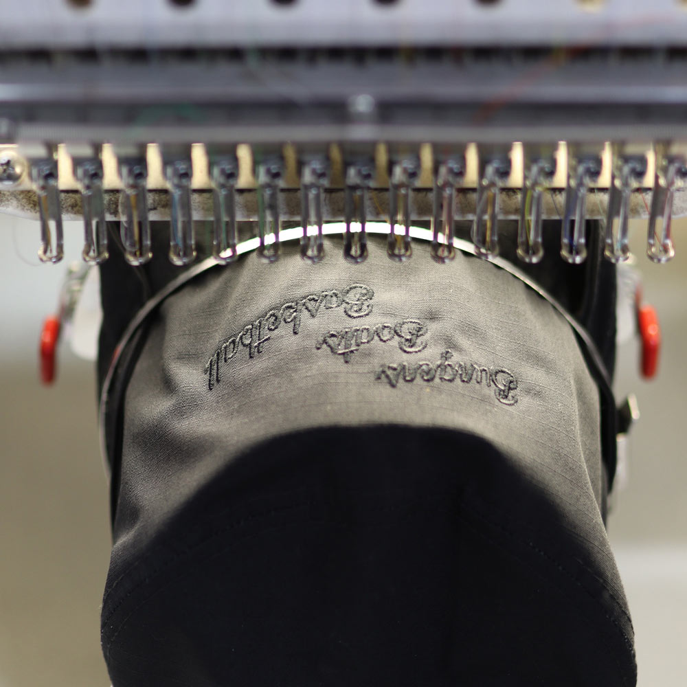 This image showcases the mass production process of the Decky 450 Fisherman’s Hat under a Barudan embroidery machine. Rows of Pigment Black hats are being meticulously embroidered with the tonal "Burger Beats Basketball" design, highlighting Garment Decor’s ability to deliver consistent quality at scale. The advanced Barudan equipment ensures flawless detailing in every stitch, reflecting Garment Decor’s commitment to excellence in custom embroidery.