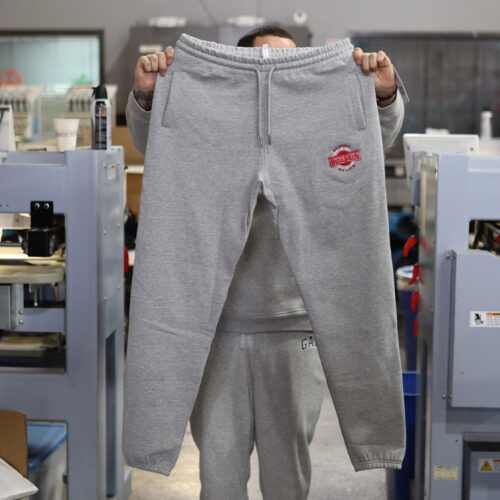 A man displays the front view of the Original Favorites Heather Grey GOTS® Organic Cotton Sweatpants, showcasing the custom embroidery in vibrant red and white thread. The bold embroidery stands out beautifully against the soft heather grey organic cotton fabric, combining eco-conscious materials with expert craftsmanship. Designed and embroidered by Garment Decor, these sweatpants redefine sustainable style and personalized branding.