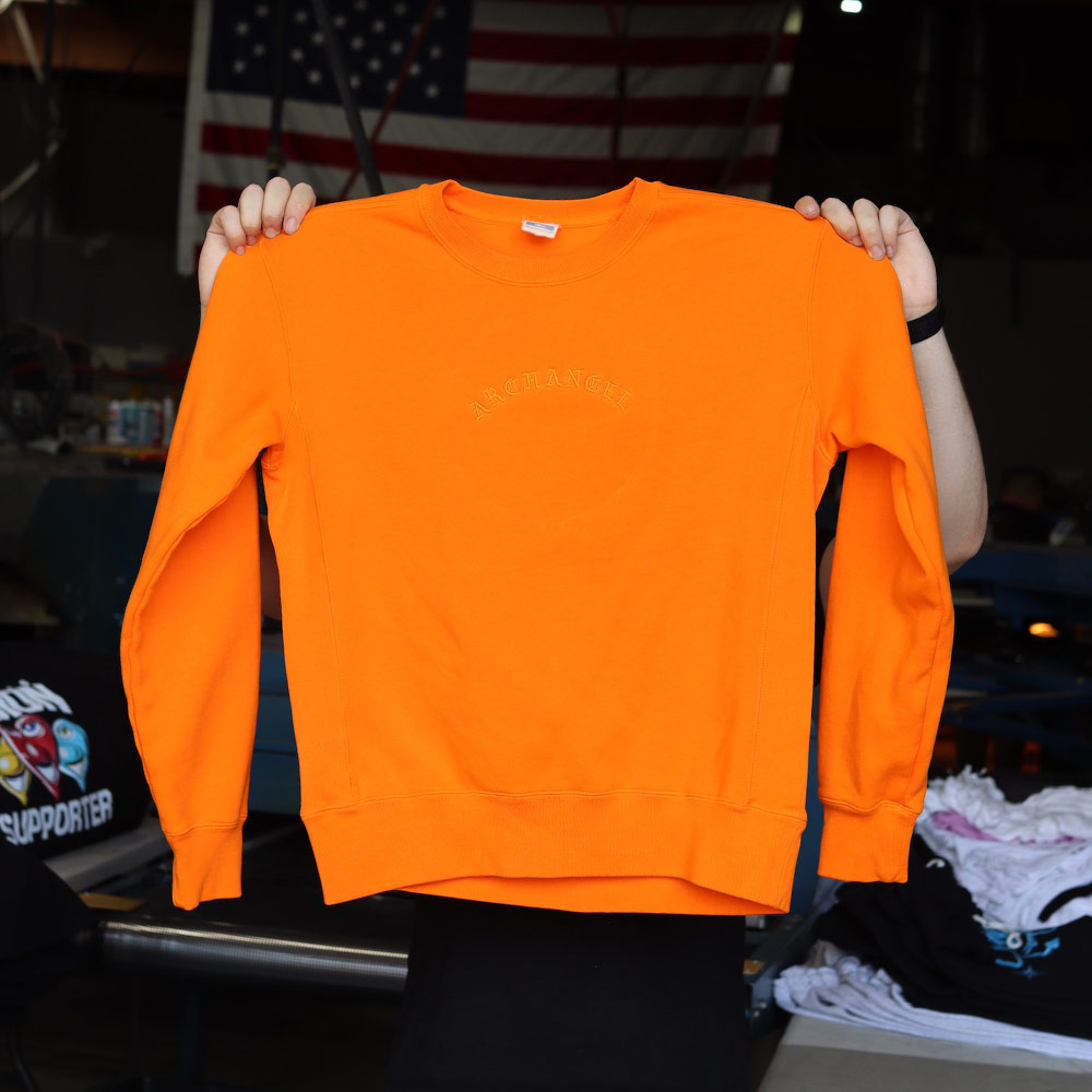 Made Blanks Varsity Crewneck Bright Orange Custom Tonal Embroidery with Arch Angel design embroidered by Garment Decor zoomed front view - Garment Decor A man displaying the front of the Made Blanks Varsity Crewneck in bright orange, showcasing the tonal embroidery for logo 'Arch Angel' design embroidered by Garment Decor. Perfect for bold, standout style.
