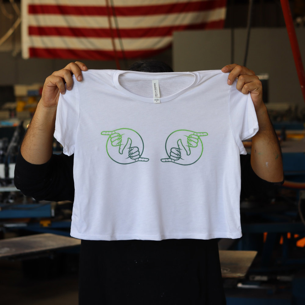 A man holding the front view of Bella + Canvas 8882 Women’s Flowy Crop Tee in white, featuring the playful 'Boob Shaka Laka' ombre design. Custom screen-printed by Garment Decor, this tee combines vibrant artistry with a modern silhouette.