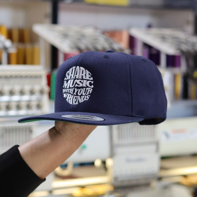 A man holds the front view of Flexfit 6477 Wool-Blend Cap in Navy, highlighting the intricate custom embroidery of the 'Share Music with Your Friends' design. Professionally embroidered by Garment Decor, this premium cap combines comfort and creativity for a standout look.