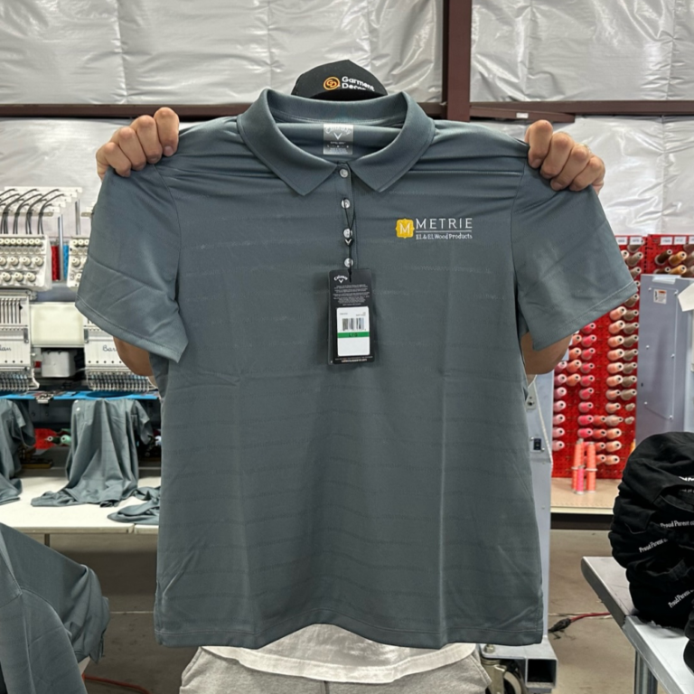 This is a photo of a gray polo that Garment Decor custom embroidered with a logo on the left chest and is being held up by our embroidery operator.