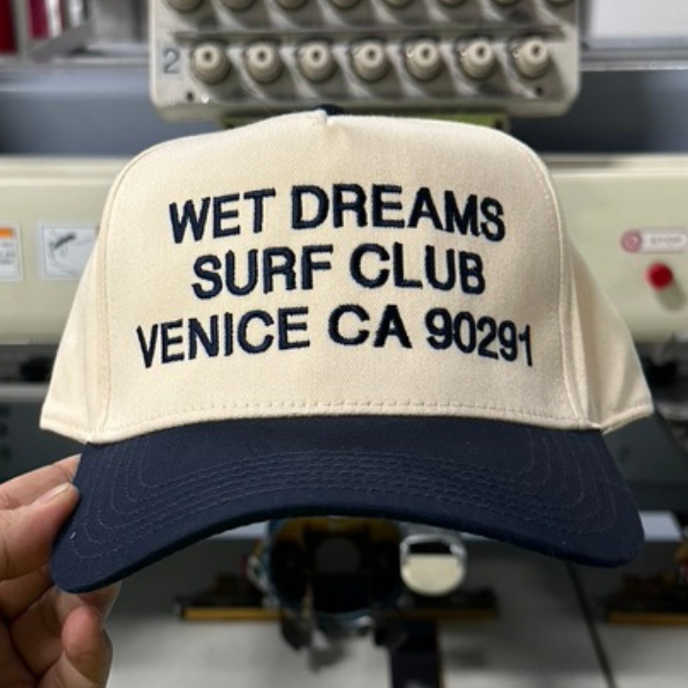 OTTO CAP 31-069 5 Panel Mid Profile Baseball Cap Navy/Natural Custom Embroidery Wet Dreams Surf Club Venice CA 90291 design embroidered by Garment Decor front view - Garment Decor A man holds the front view of OTTO CAP 31-069 5 Panel Mid Profile Baseball Cap in a striking Navy/Natural colorway, featuring custom embroidery of the "Wet Dreams Surf Club Venice CA 90291" design. The embroidery showcases a bold, vintage-inspired script for "Wet Dreams Surf Club," elegantly curved to evoke a retro surf culture vibe. Below, "Venice CA 90291" is embroidered in a clean, understated font, adding a sense of authenticity and locality to the design. The combination of vibrant navy panels and a natural-colored bill enhances the cap's classic aesthetic, while the high-quality, textured embroidery by Garment Decor ensures a durable and premium finish.