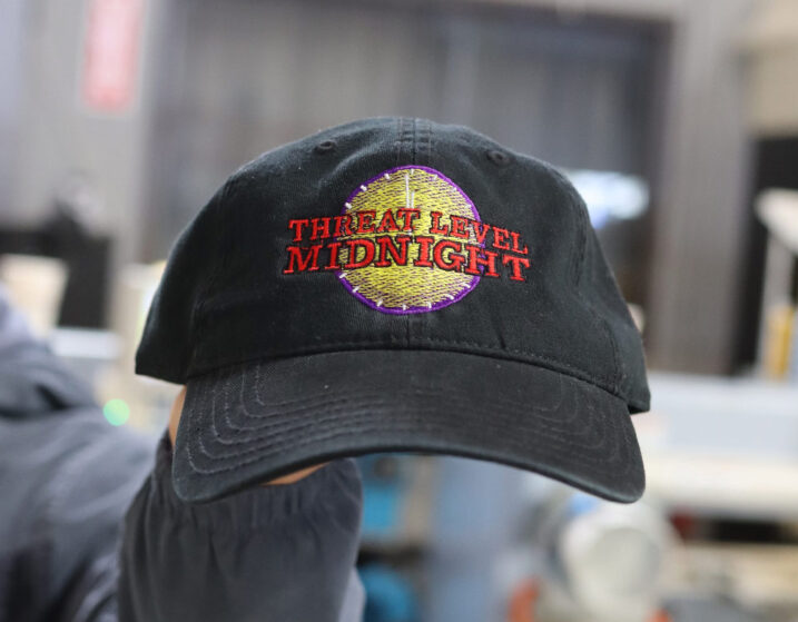 A black baseball cap with the words "threat level midnight" embroidered in red and a multi-colored design in the background.