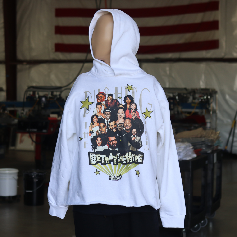 Person wearing a white hoodie with a graphic print of a group of people on the back, standing in an industrial or workshop setting with an american flag in the background.