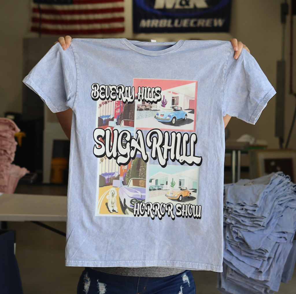 Person holding up a blue t-shirt with a graphic depicting various scenes and the text "beverly hues, sugarhill, horror show" printed on it.