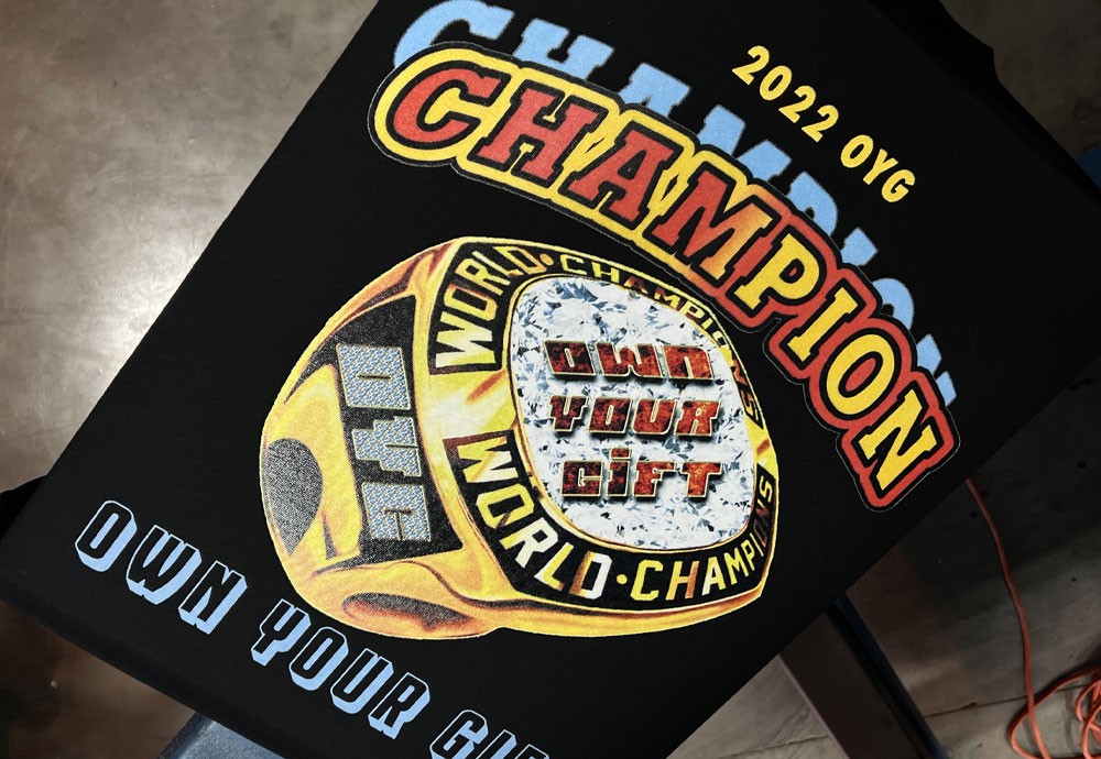 Black t-shirt with a colorful "2022 vg champion" graphic design featuring phrases "own your world-champion" and "world champion" with a stylized golden trophy.