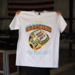 A person holding up a white t-shirt with a colorful "champion own your gift" graphic design.