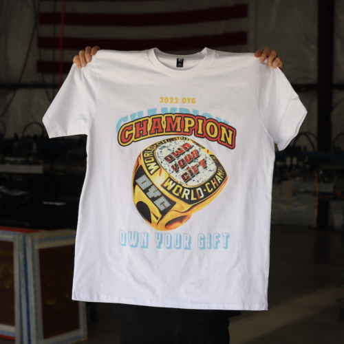 A person holding up a white t-shirt with a colorful "champion own your gift" graphic design.