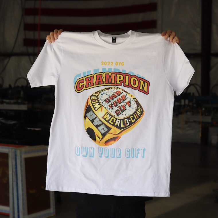 A person holding up a white t-shirt with a colorful "champion own your gift" graphic design.