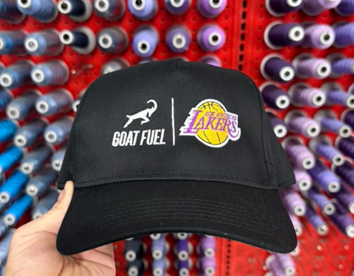 A man holds the OTTO CAP 31-069 5 Panel Mid Profile Baseball Cap in sleek black, showcasing its custom embroidery in the front view. The design features the Goat Fuel logo embroidered on the right side and the iconic LA Lakers logo on the left, presented side by side for a balanced and striking visual impact. The high-quality threadwork ensures crisp details and vibrant colors, emphasizing both logos' professional and premium look, embroidered by Garment Decor.