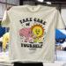 A person holding up a beige t-shirt with a graphic design that says "take care of yourself" and features illustrated characters, inside a workshop setting.