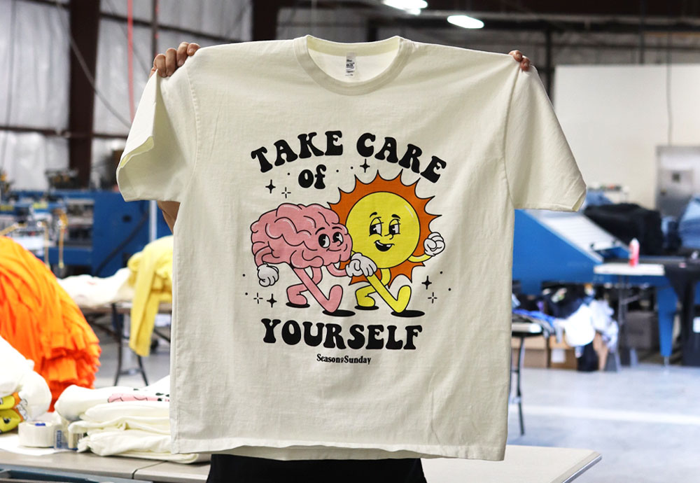 A person holding up a beige t-shirt with a graphic design that says "take care of yourself" and features illustrated characters, inside a workshop setting.