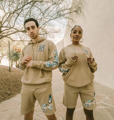 Two individuals modeling matching beige hoodies and shorts with blue graphic designs for brands.