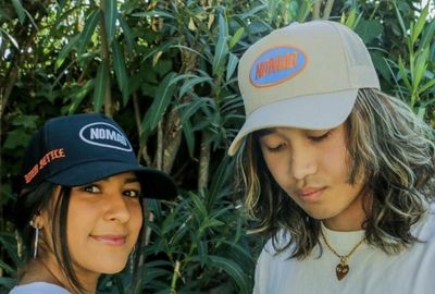 Two individuals wearing baseball caps with logos embroidered for brands stand in front of green foliage.