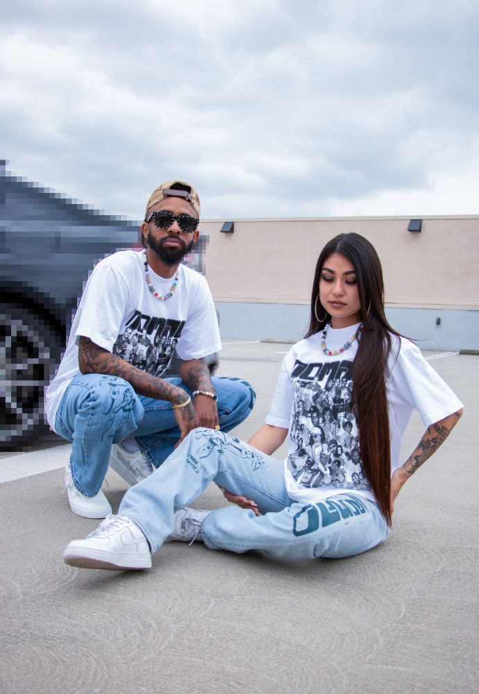 Two people wearing graphic tees and jeans sitting on concrete with a cloudy sky in the background.