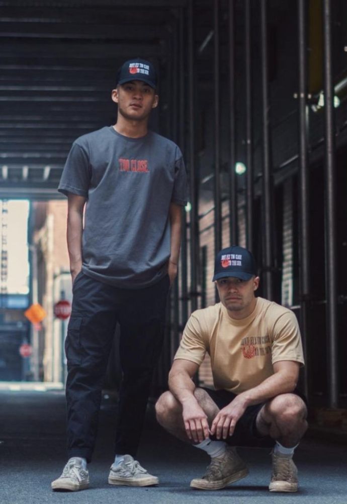 Two men wearing graphic t-shirts and baseball caps standing in an urban alleyway.