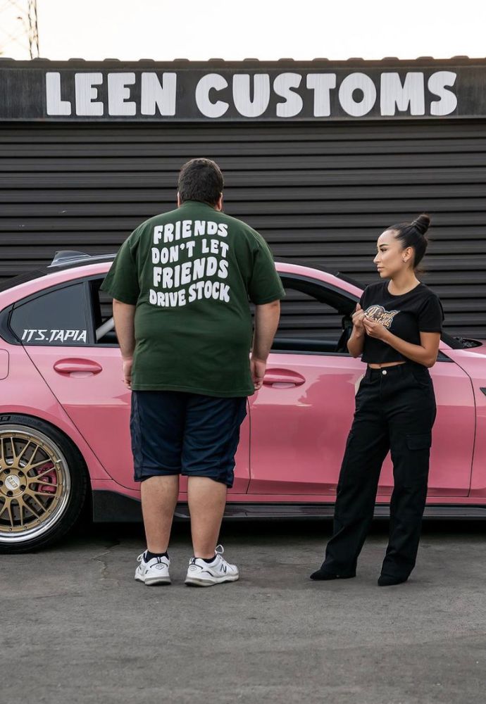 Two people standing by a customized pink car, with the man wearing a t-shirt that reads "friends don't let friends drive stock".