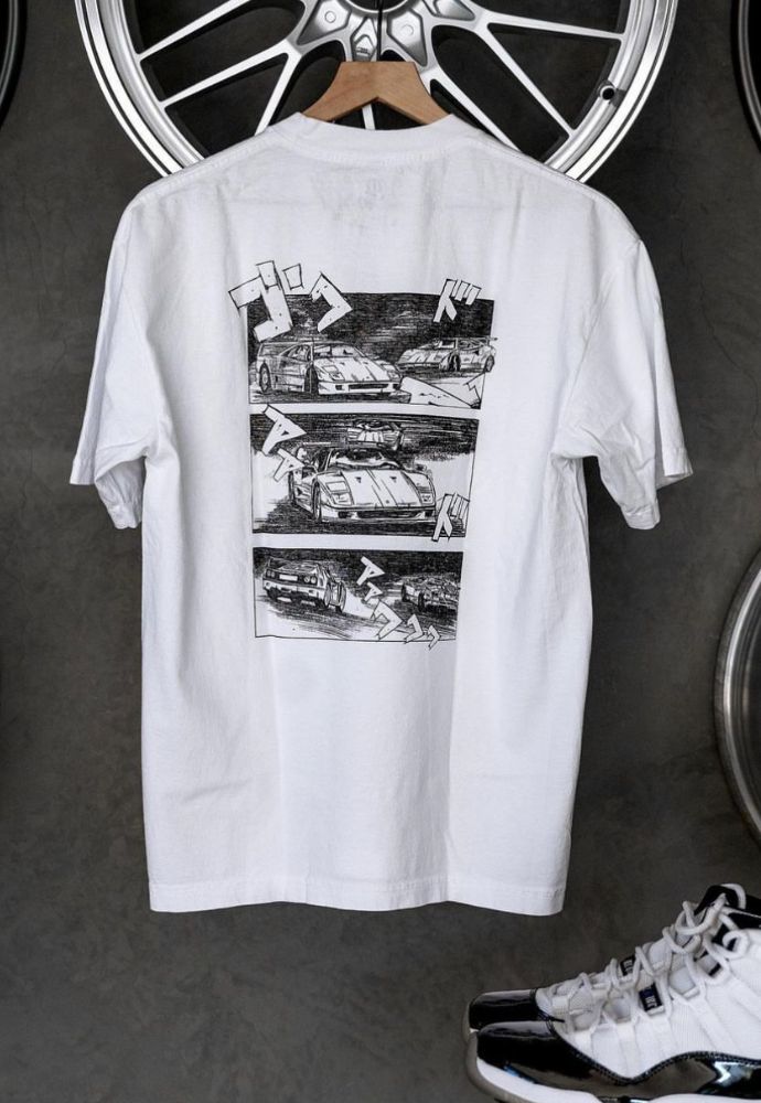 White t-shirt with graphic print hanging against a gray background flanked by wheel rims and sneakers.